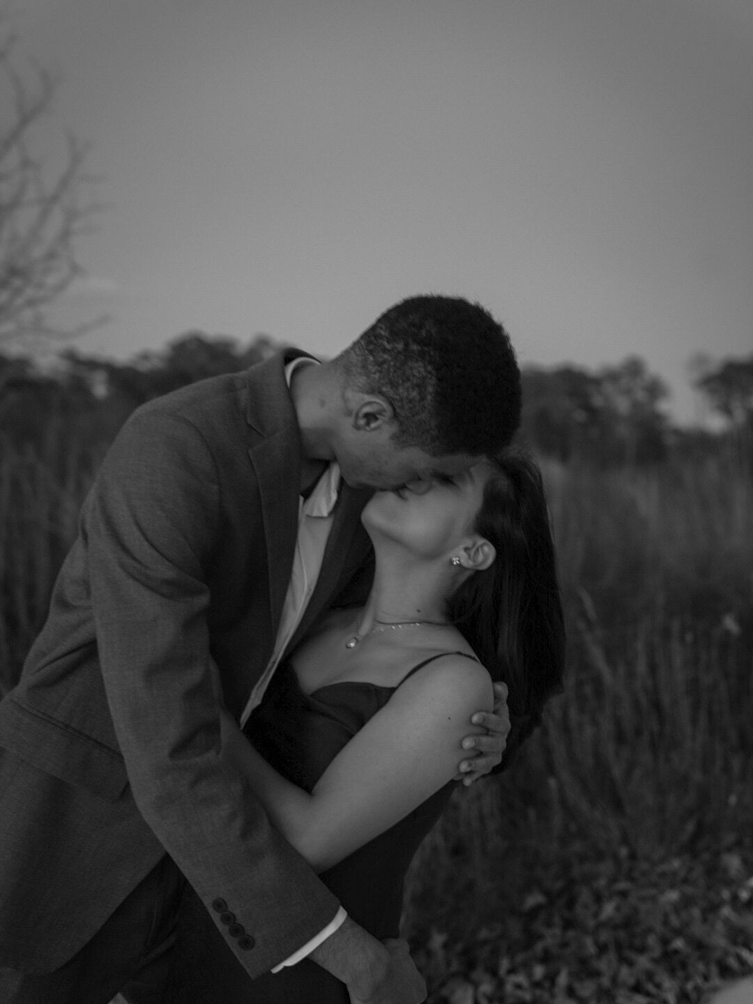Black and white photo of a couple sharing a romantic kiss outdoors at dusk.