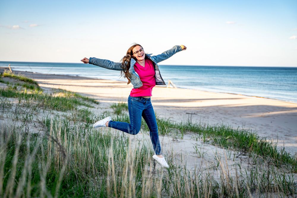 Young woman joyfully jumping on a sandy beach with arms outstretched, wearing a pink top and denim jacket.
