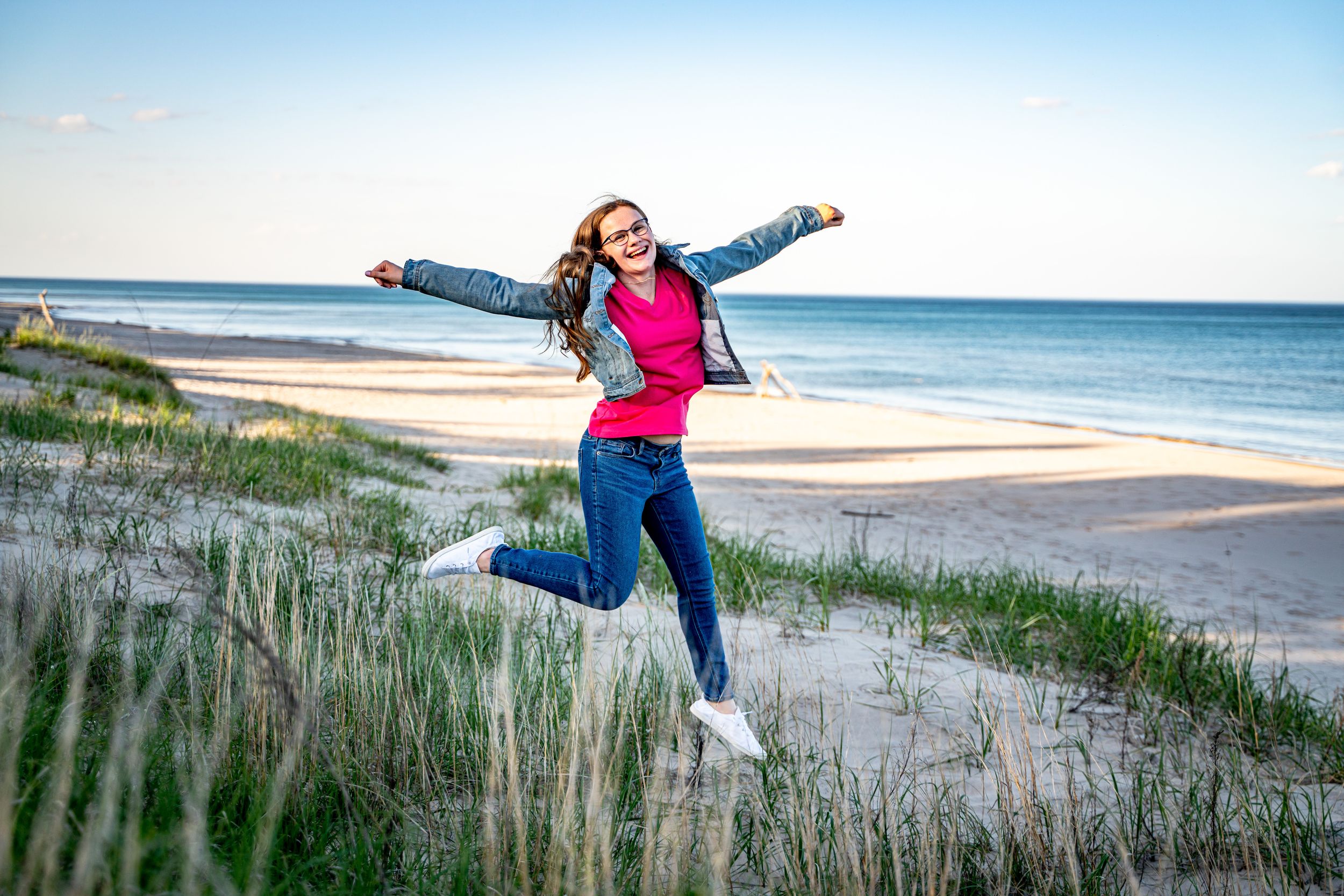 Young woman joyfully jumping on a sandy beach with arms outstretched, wearing a pink top and denim jacket.
