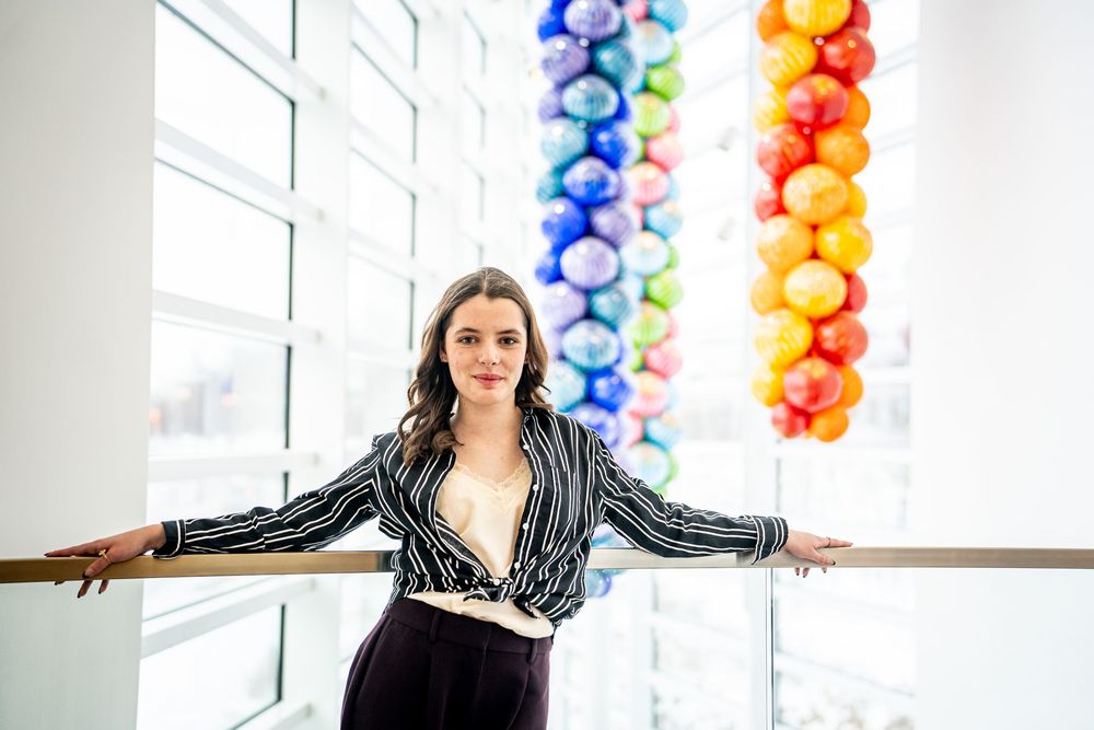 Young woman in striped blazer poses near colorful glass ball art installation indoors.