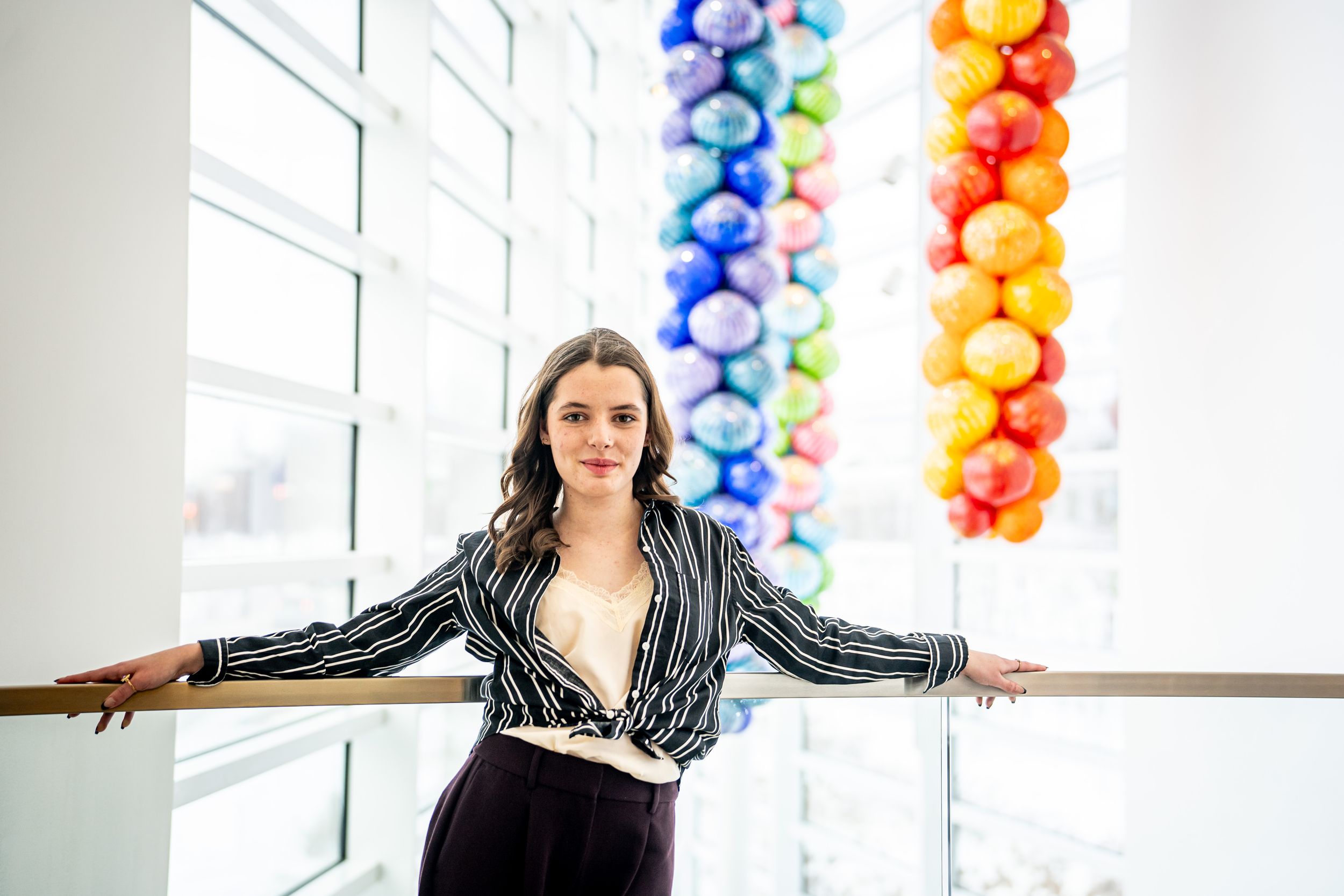 Young woman in striped blazer poses near colorful glass ball art installation indoors.