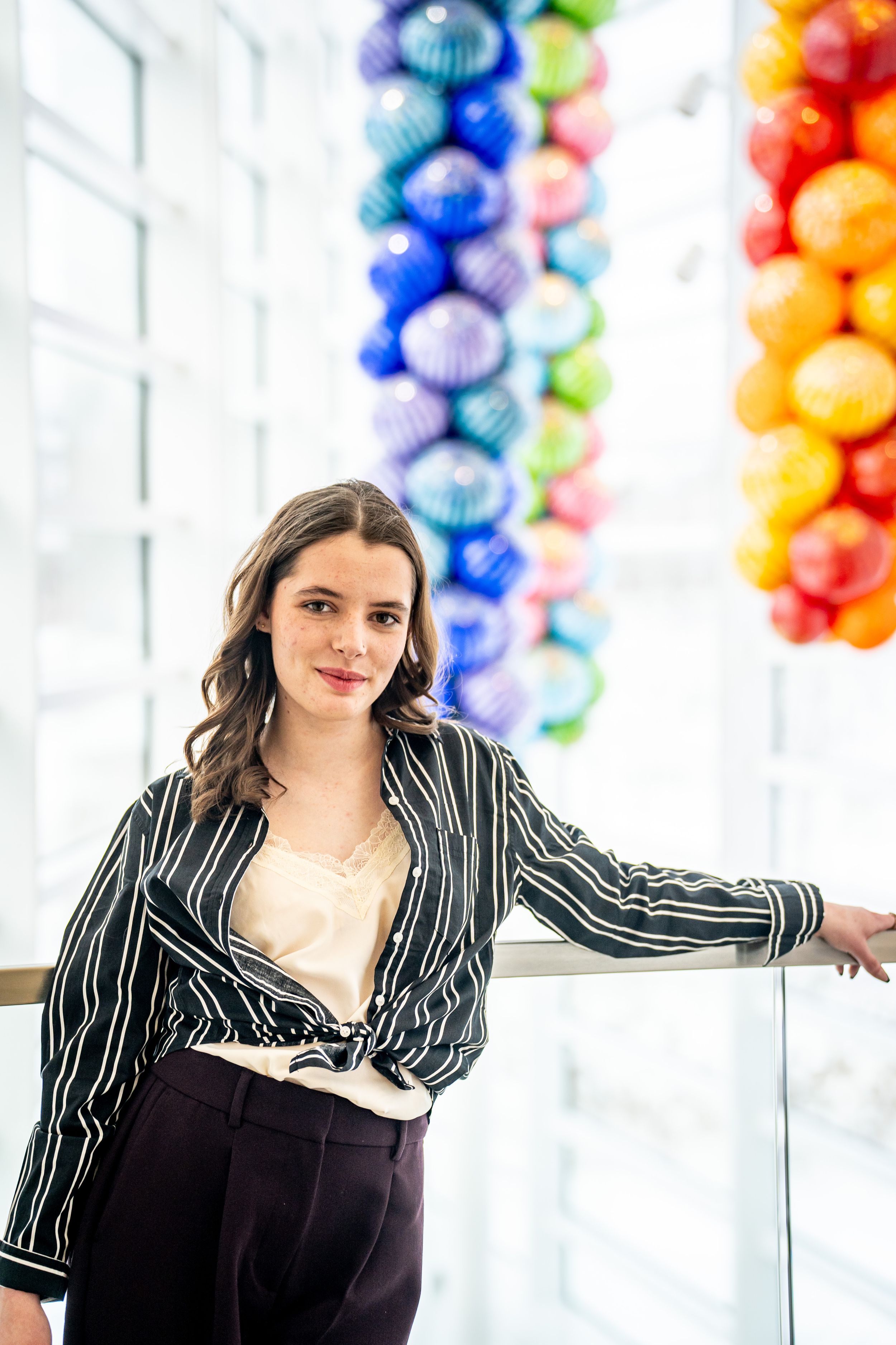 Young woman in striped blouse poses near railing with colorful glass ball art installation in bright modern building.