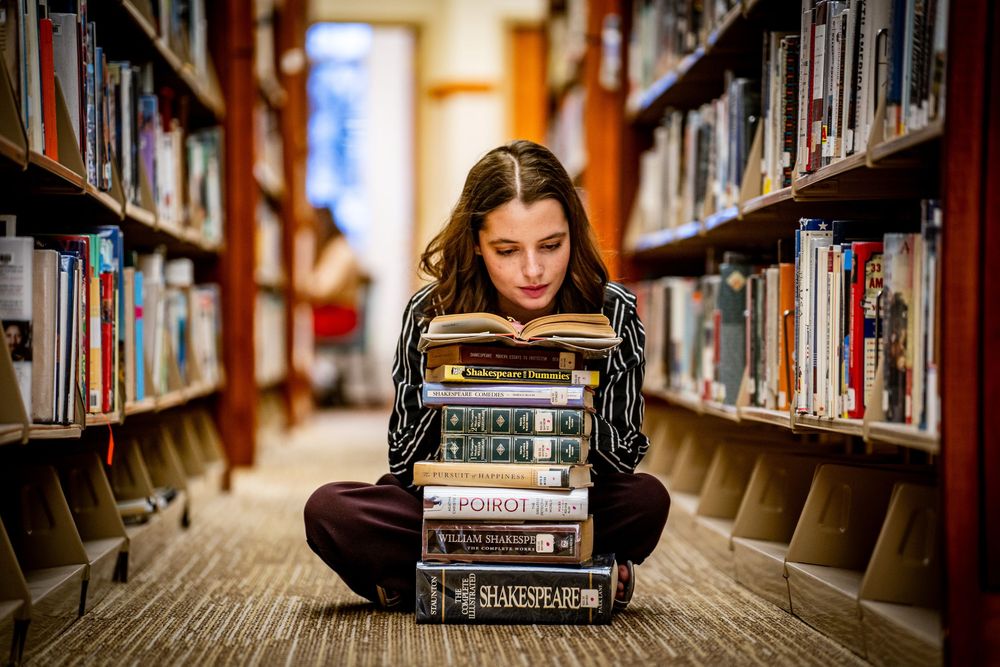 Girl sits cross-legged on library floor reading from a tall stack of classic books.