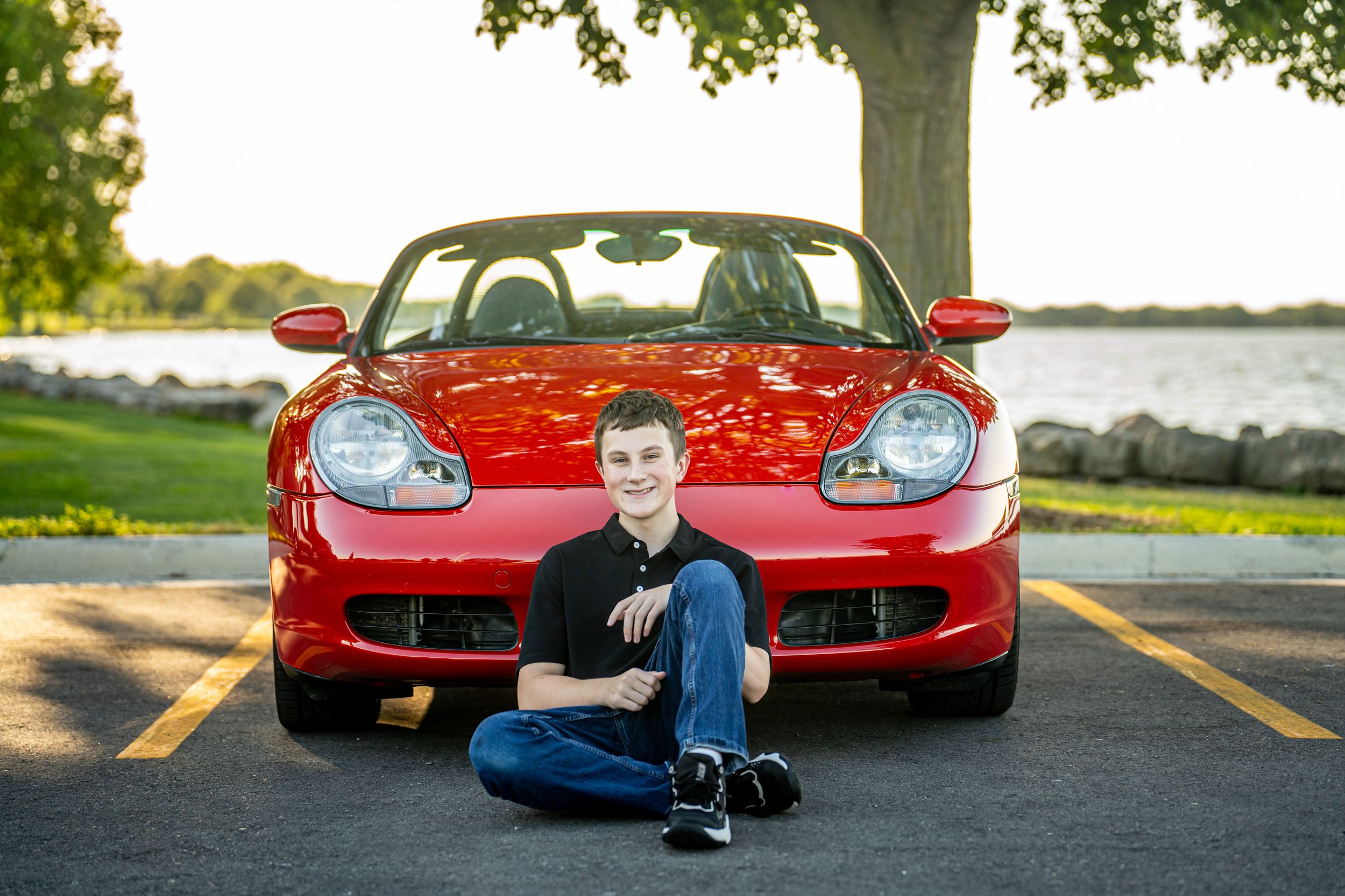 Young man sitting cross-legged in front of a red Porsche Boxster convertible in a scenic waterfront parking lot. senior portrait