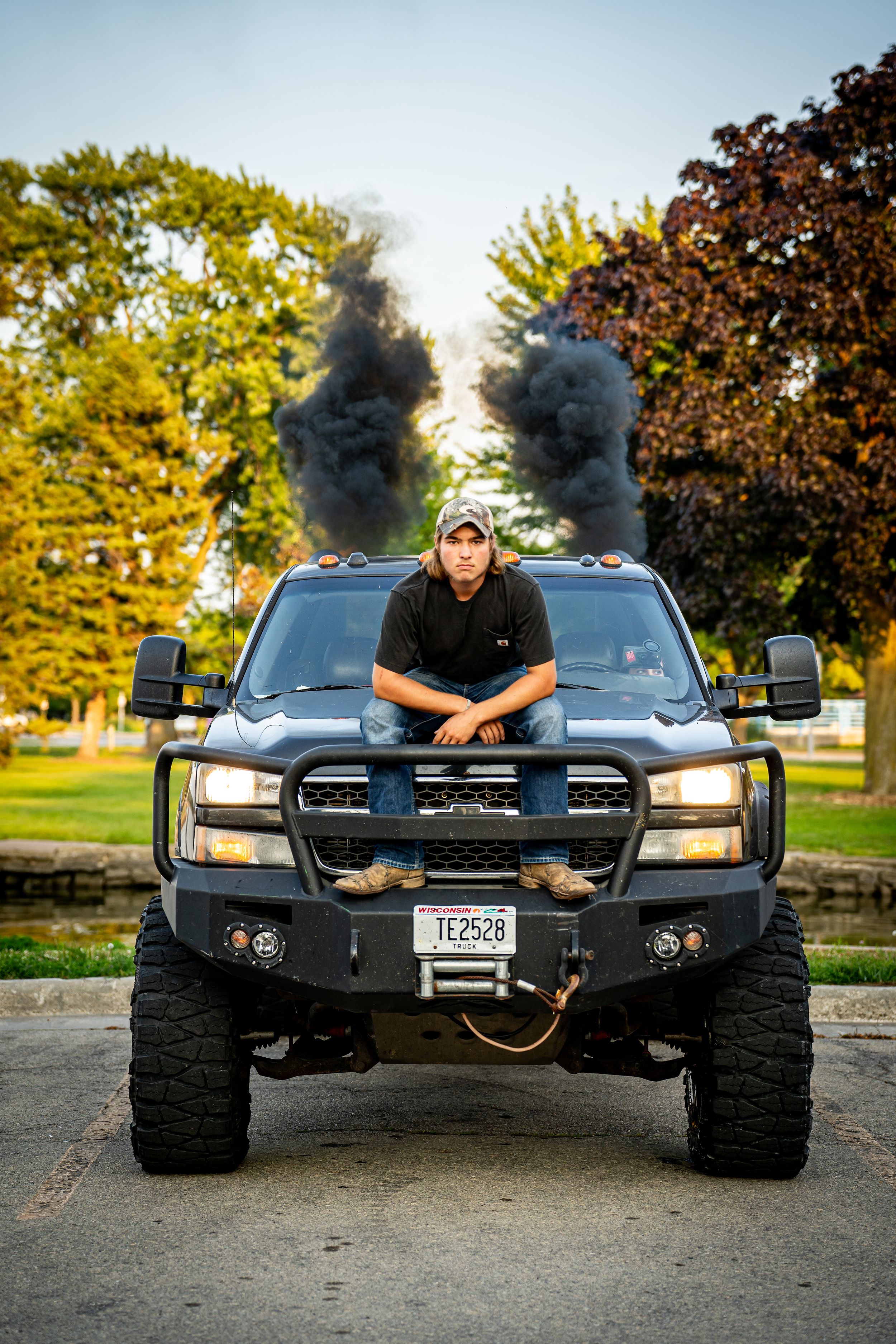 Young man poses on lifted black diesel truck emitting dark smoke from dual exhaust stacks outdoors.
