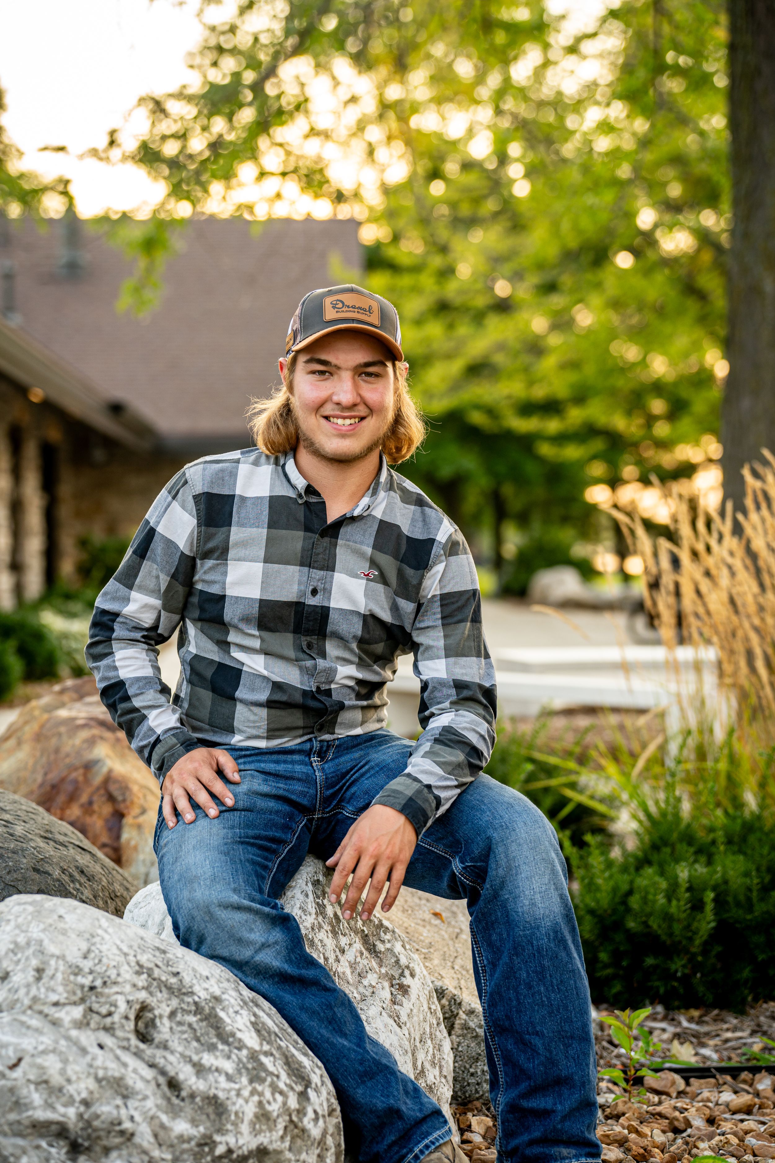 Smiling teen in plaid shirt and cap sits on rocks in a lush green garden setting.