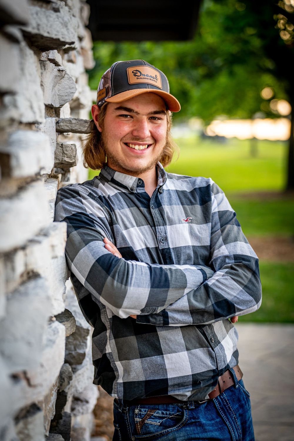 Smiling young man in plaid shirt and baseball cap leaning against stone wall outdoors, posing for a senior photoshoot