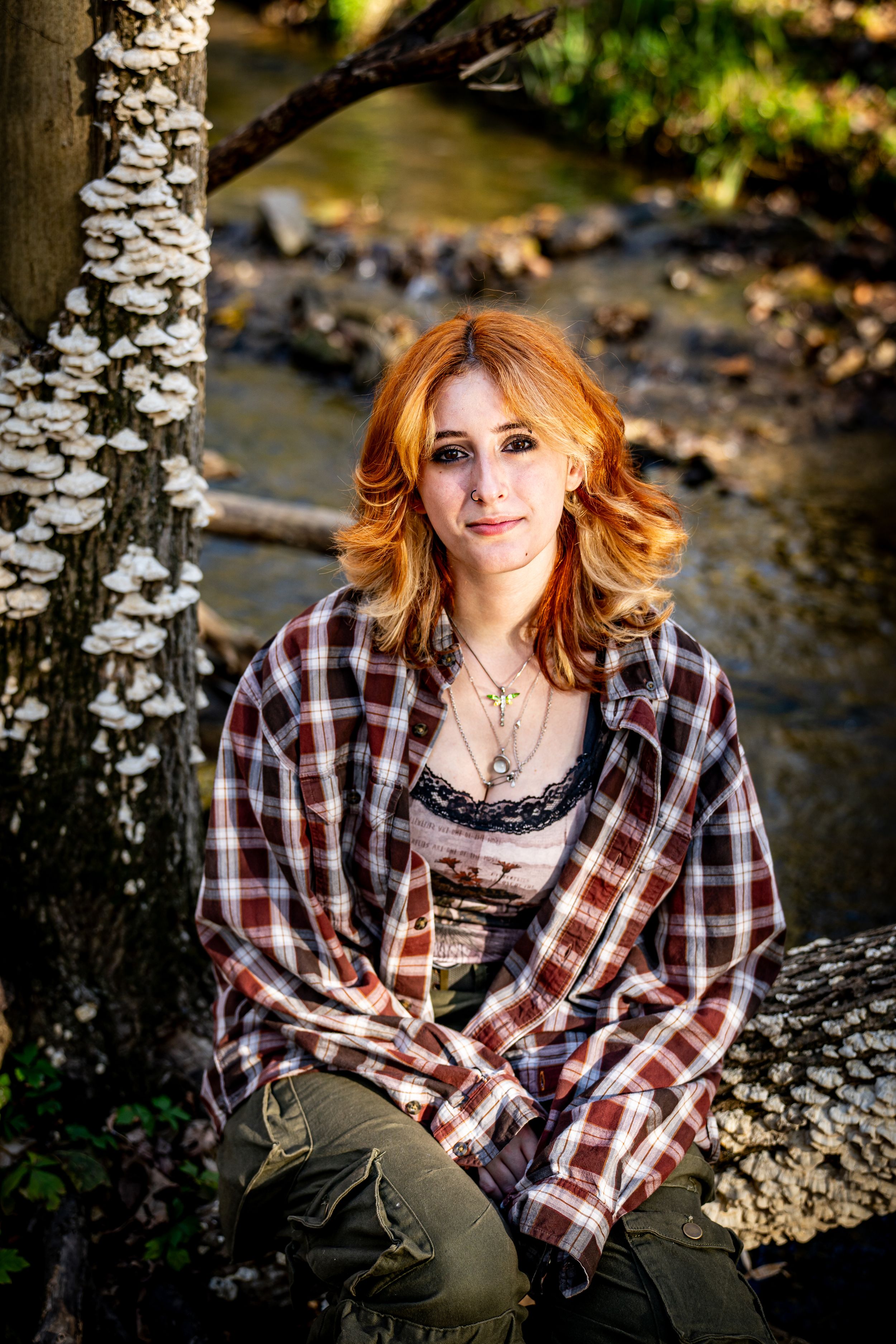 Red-haired woman in plaid shirt sitting near a creek by a mushroom-covered tree in a forest.