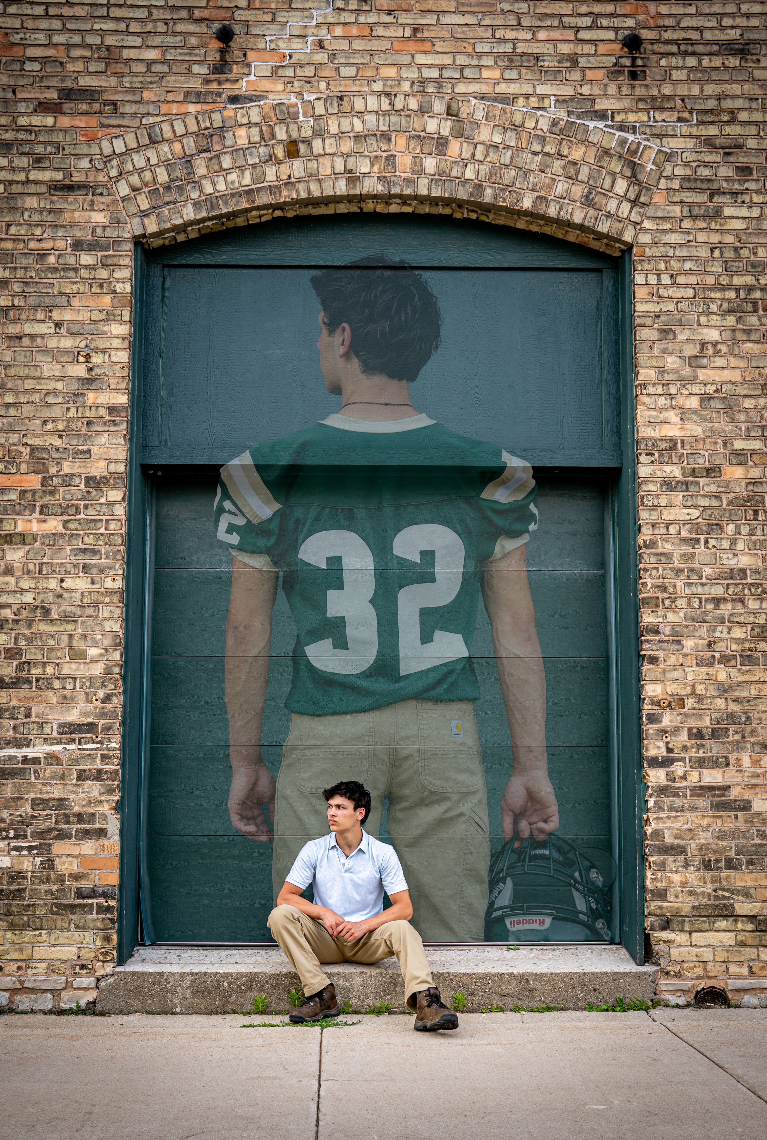 Teen boy sits before a large mural of football player number 32 on a brick wall.