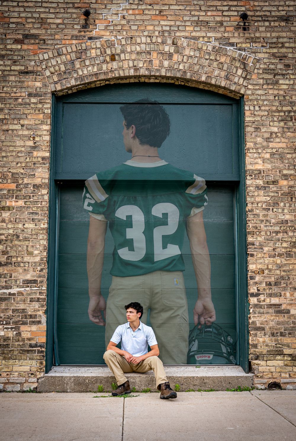 Teen boy sits before a large mural of football player number 32 on a brick wall.