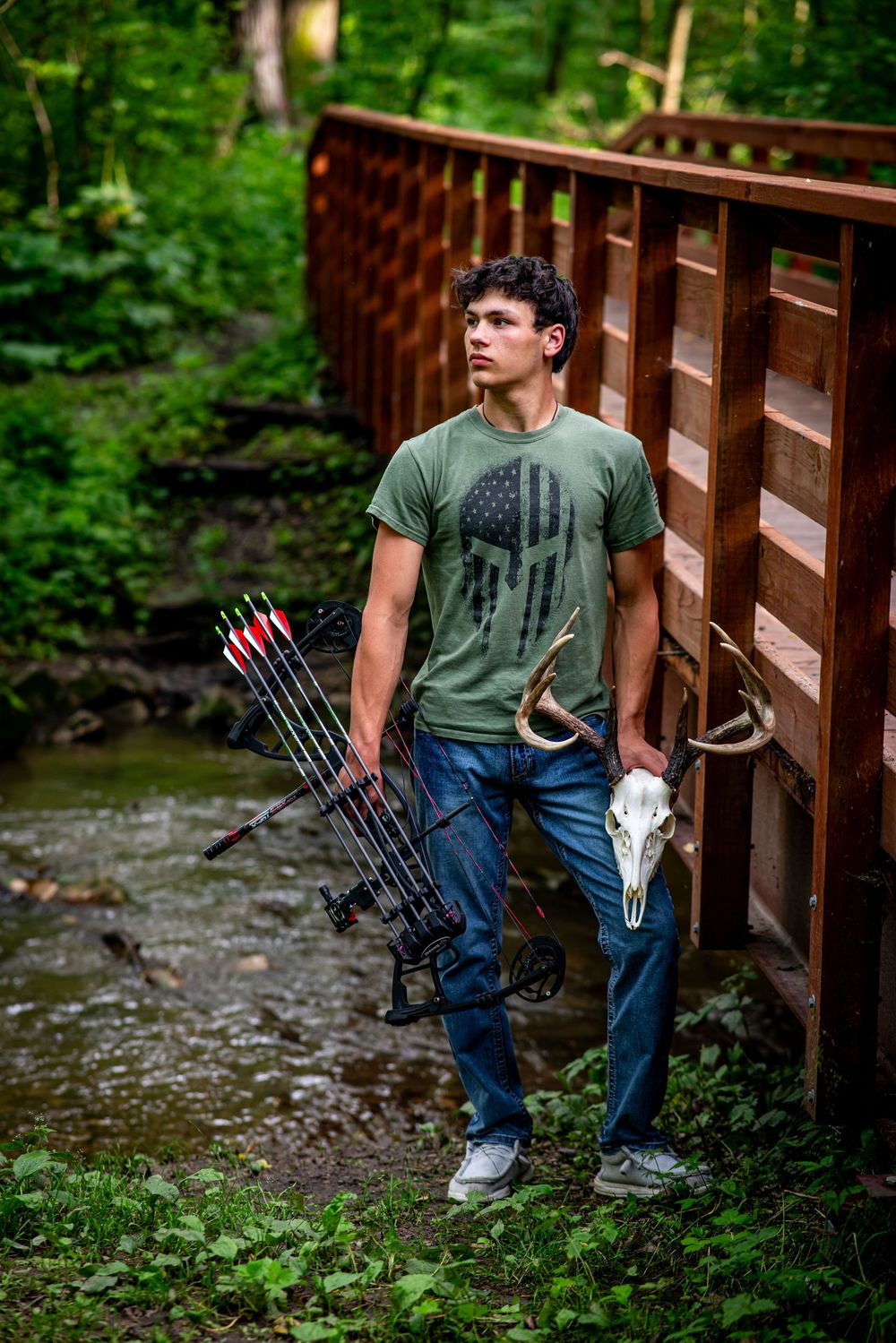 Teen boy holds compound bow and deer skull on a wooden bridge in a forest.