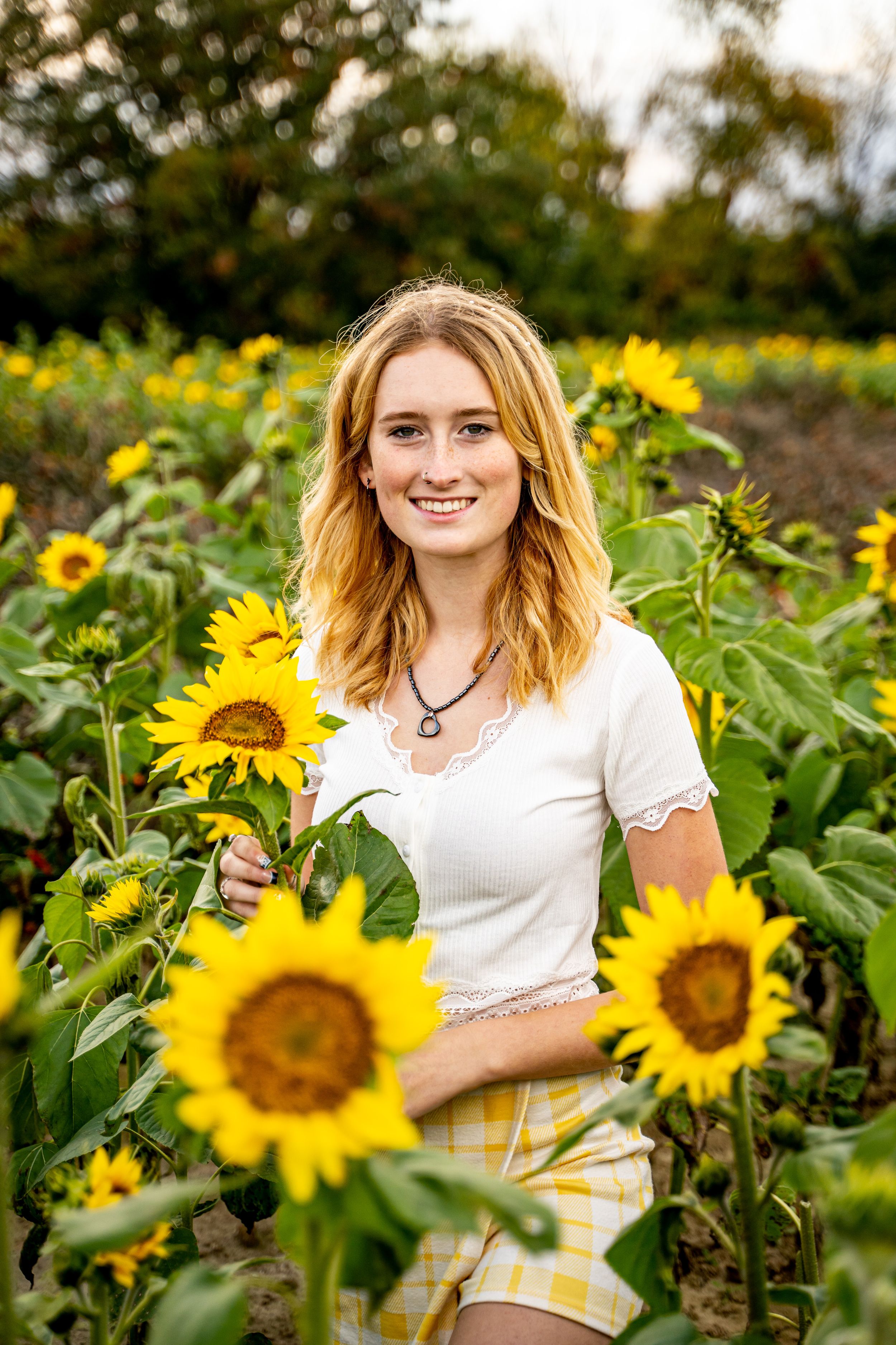 Smiling teenage girl with red hair stands among bright yellow sunflowers in a field.