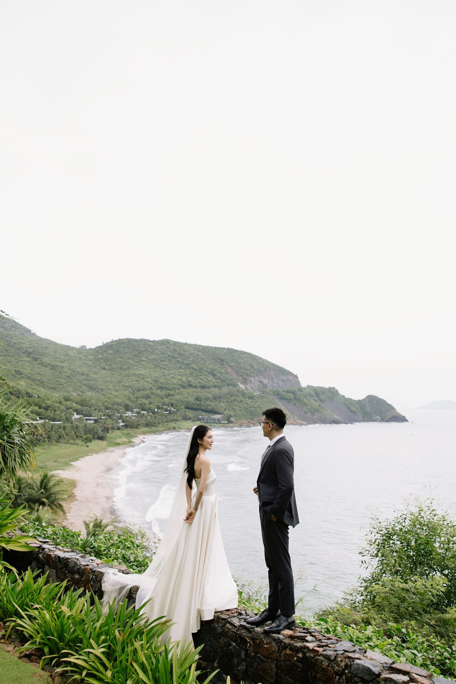 Bride and groom stand on stone wall overlooking tropical beach and green mountains on their wedding day.