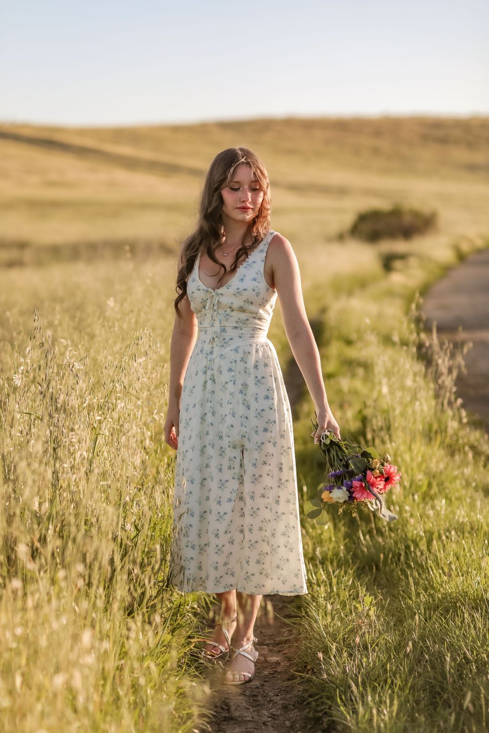 Young woman in floral midi dress walks through golden meadow holding wildflower bouquet at sunset.