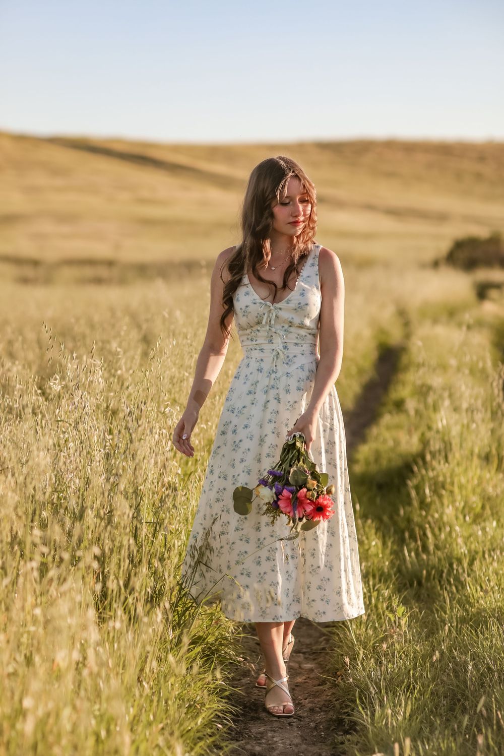 Young woman in floral midi dress walking a dirt path through golden grass fields holding wildflowers.