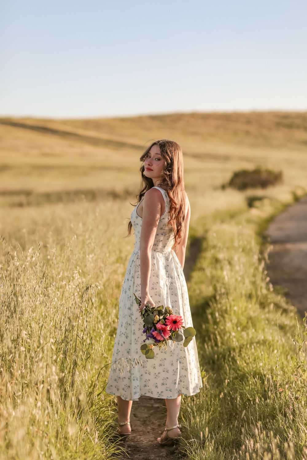 Young woman in floral dress holding pink flowers stands on a rural path surrounded by golden fields.