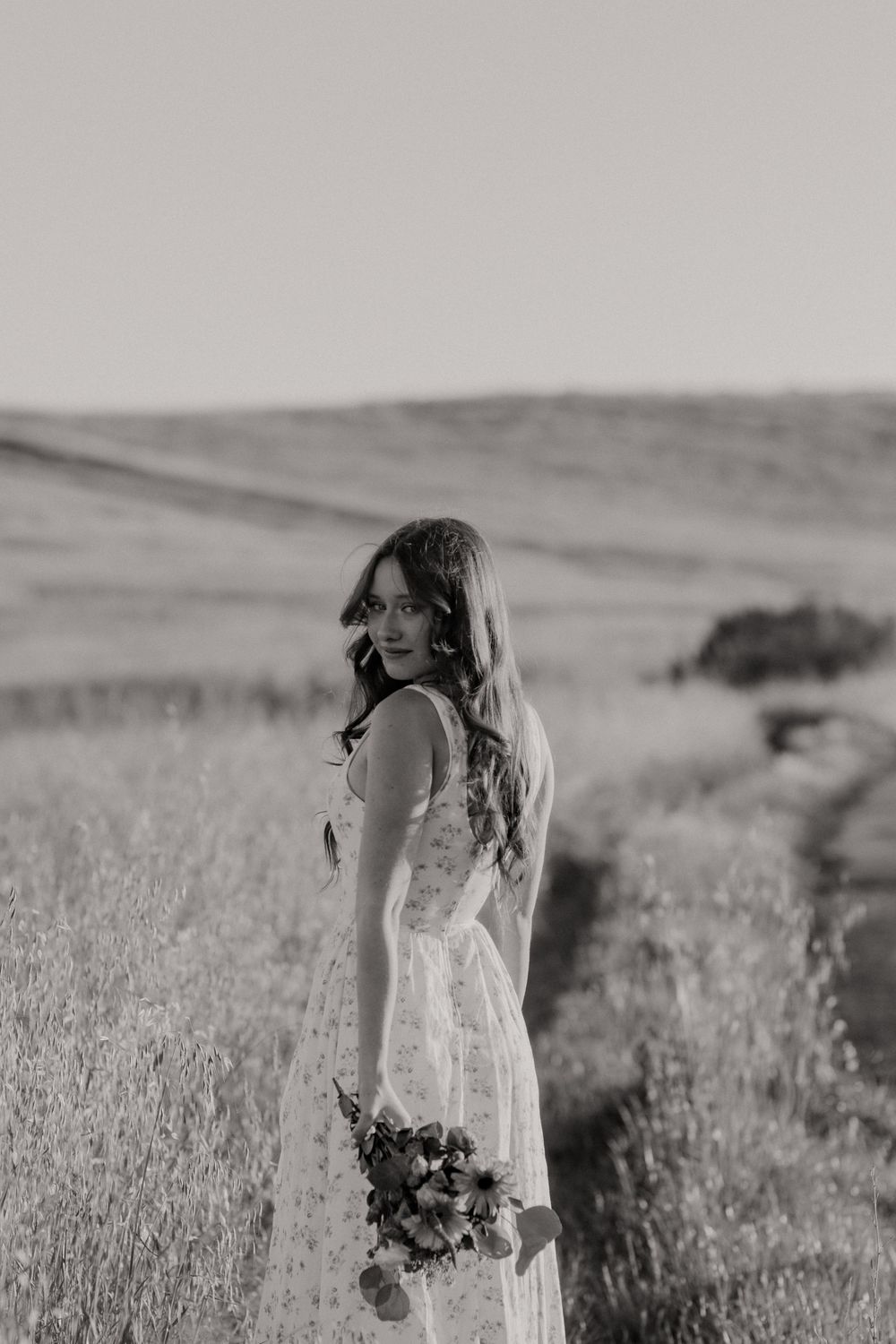 Black and white photo of a bride in a white dress holding a bouquet in a scenic field.