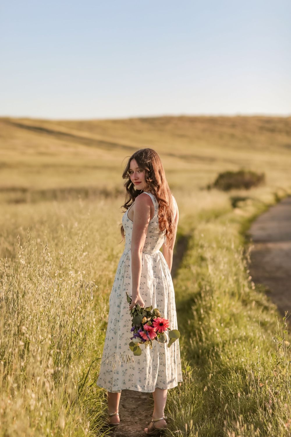 Woman in white floral sundress standing on rural path in sunlit meadow holding colorful bouquet.