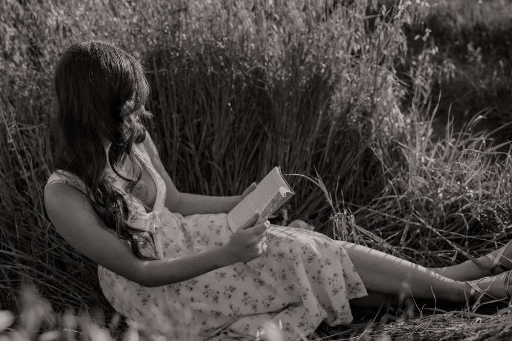 A young woman in a floral dress reads a book while sitting in a field of tall grass, in black and white.