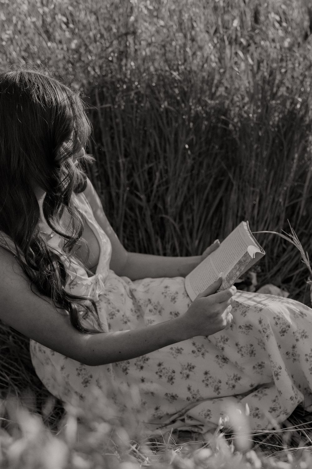 Black and white photo of a young woman in a floral dress sitting in a field, reading a book outdoors.
