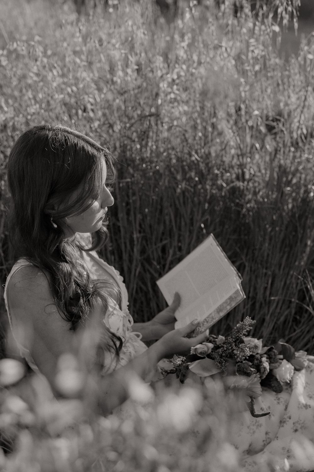 Black and white photo of a woman reading a book outdoors in a field, holding a bouquet of flowers.