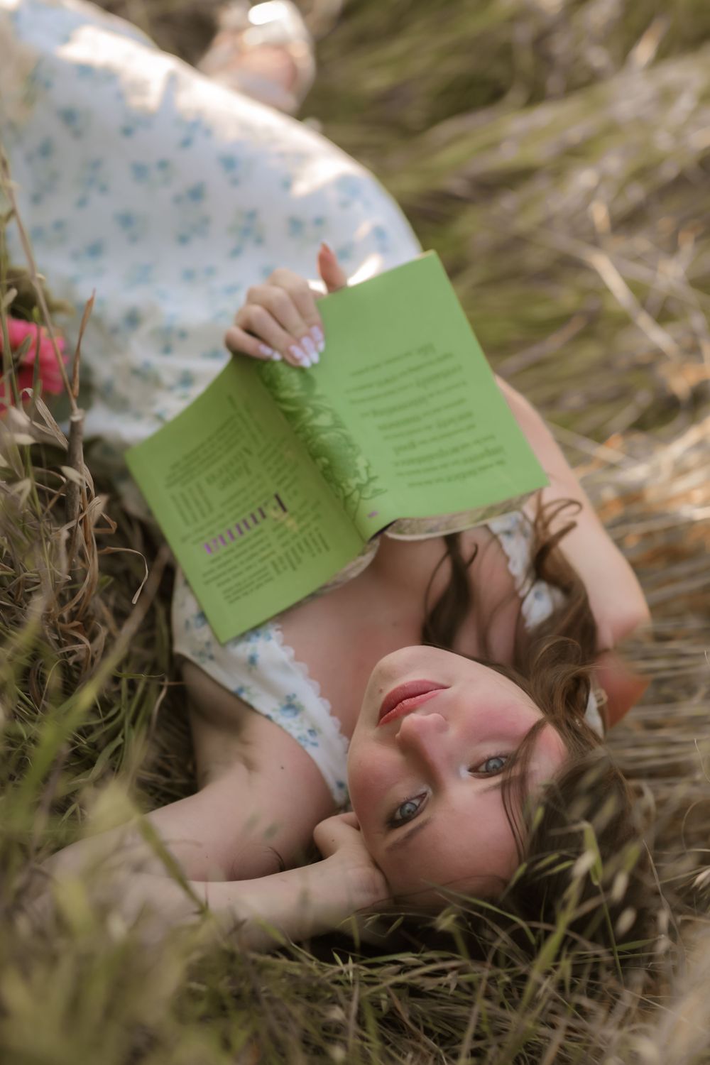Young woman lying in a field of tall grass, reading a green book outdoors on a sunny day.