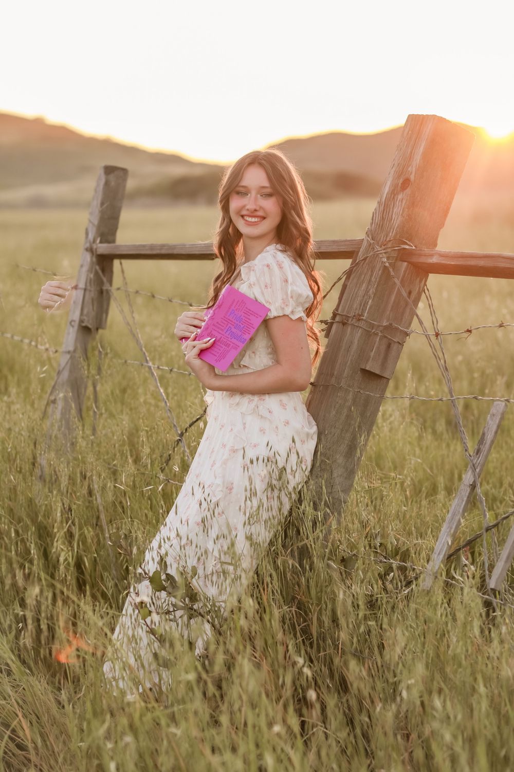 Young woman in floral dress holding pink book leaning on rustic fence in golden sunset field.
