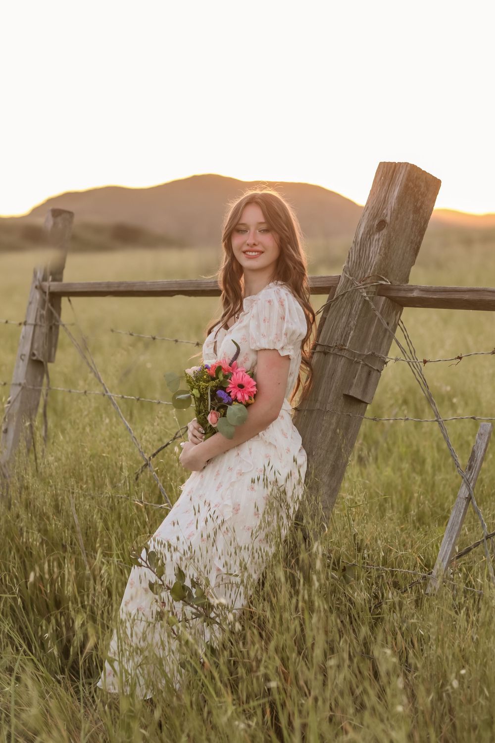 Young woman in white floral dress holding pink bouquet leans against rustic fence post in golden sunset field.