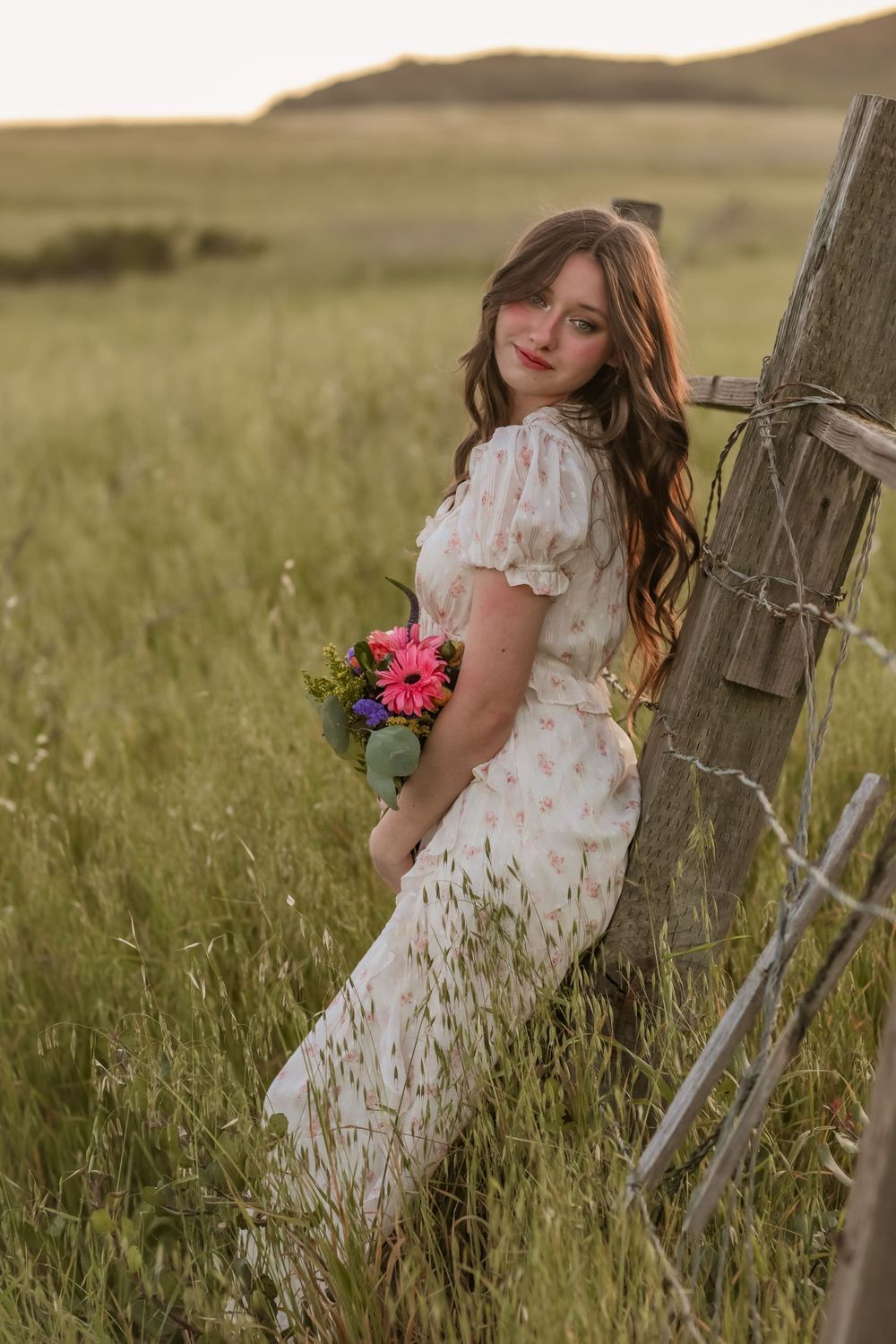 Young woman in floral dress holding wildflowers leaning on fence post in golden hour field.