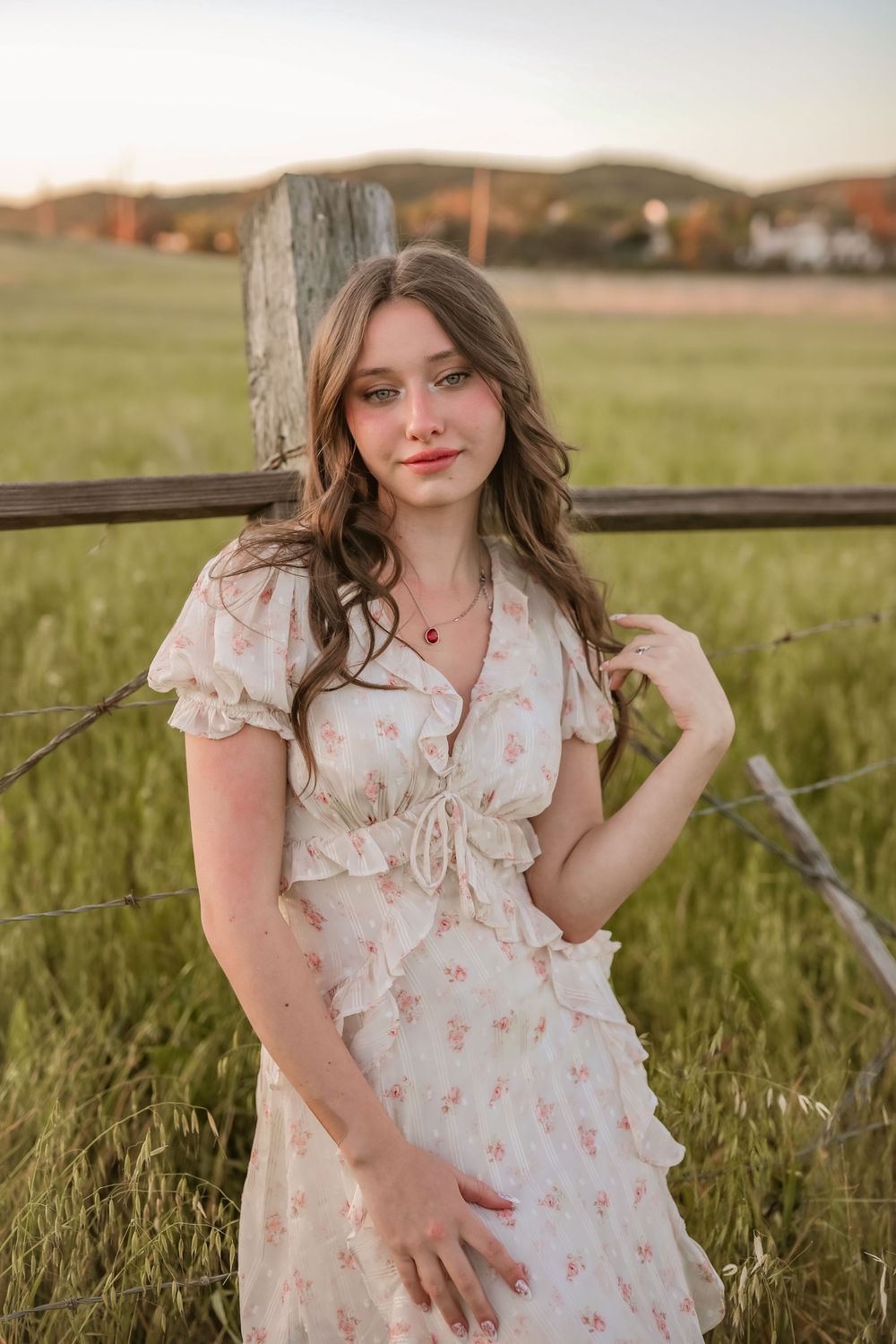Young woman in floral dress leaning against rustic fence in golden hour meadow.