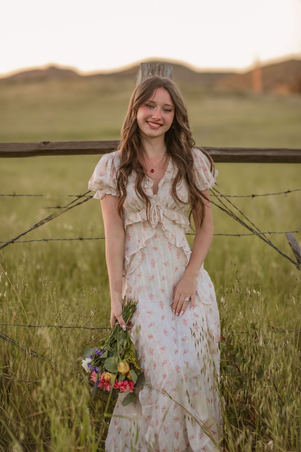 Smiling young woman holding wildflowers in floral dress at sunset fence.