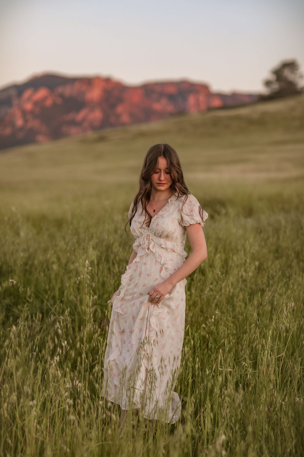 Woman in floral maxi dress standing in green field at golden hour with red rock formations in background.