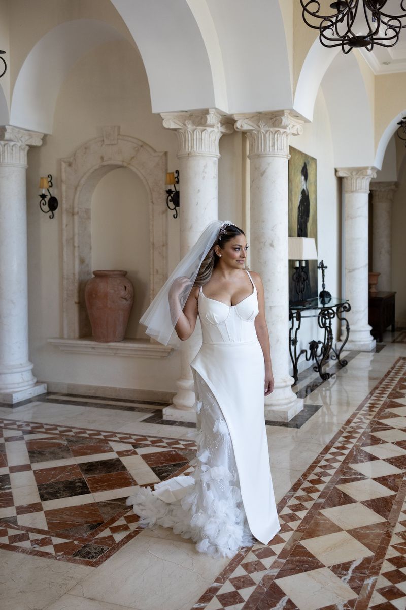 Bride in white spaghetti strap wedding gown with veil poses in elegant marble columned hall with checkered floor.