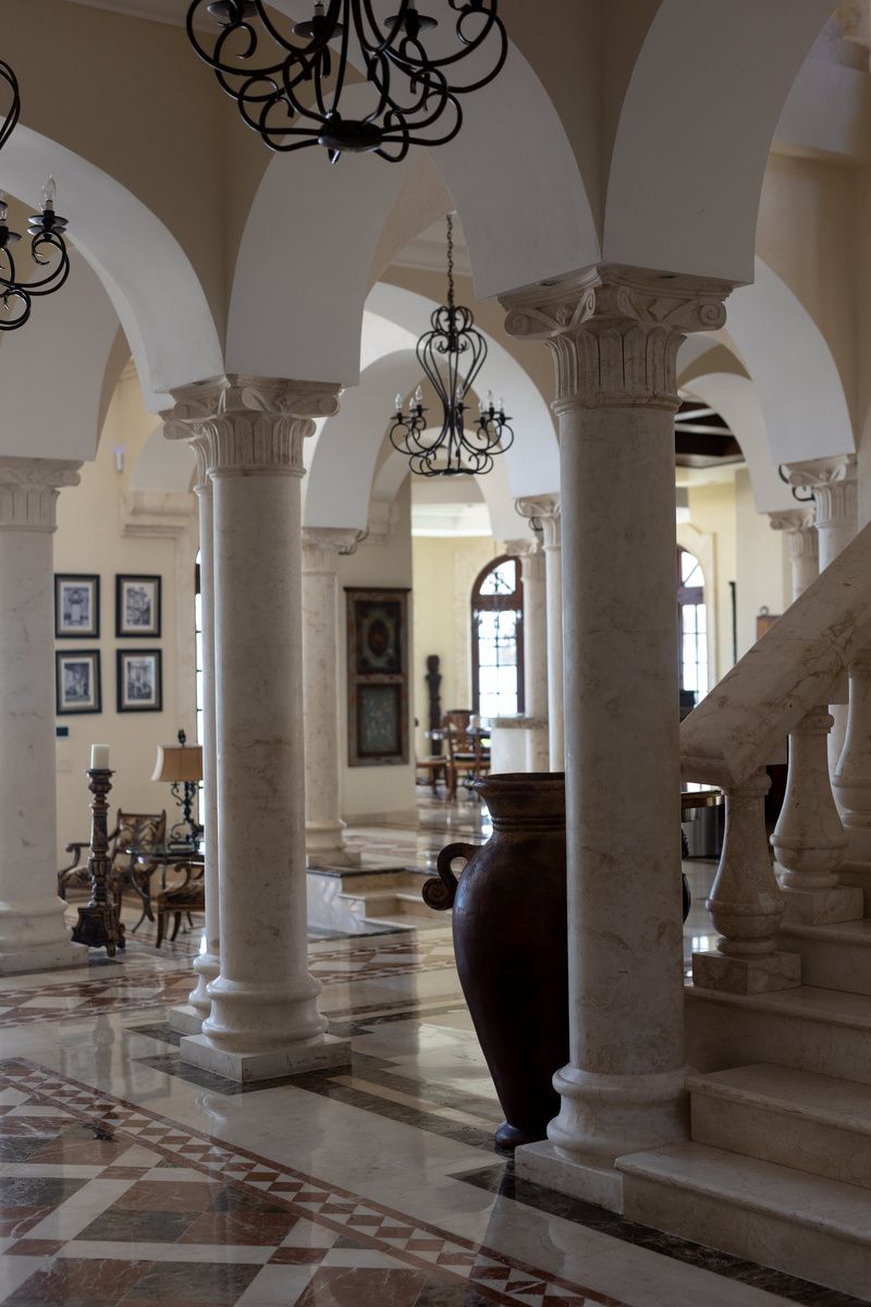 Elegant hotel lobby with marble columns, arched ceilings, ornate chandeliers, decorative vase, and grand staircase on polished floors.