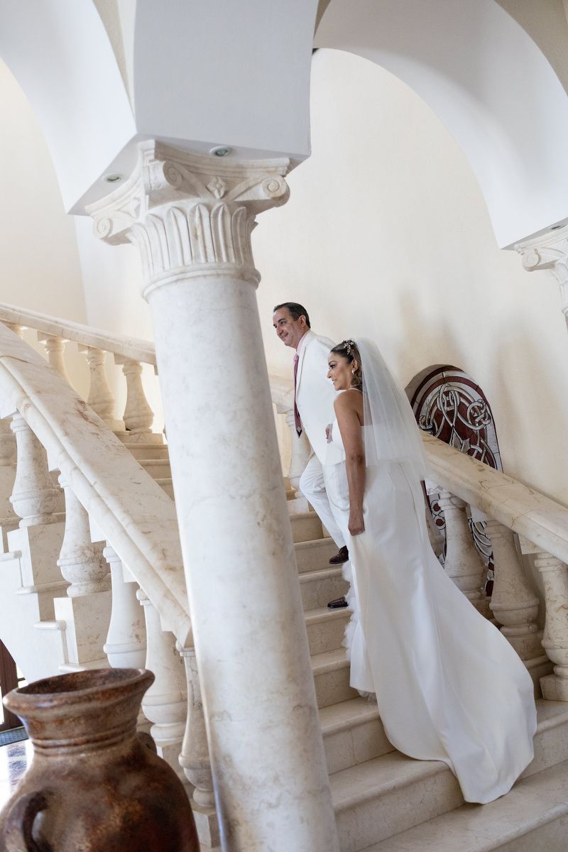 Bride and groom in white attire walking up elegant marble staircase with ornate columns in a grand venue.