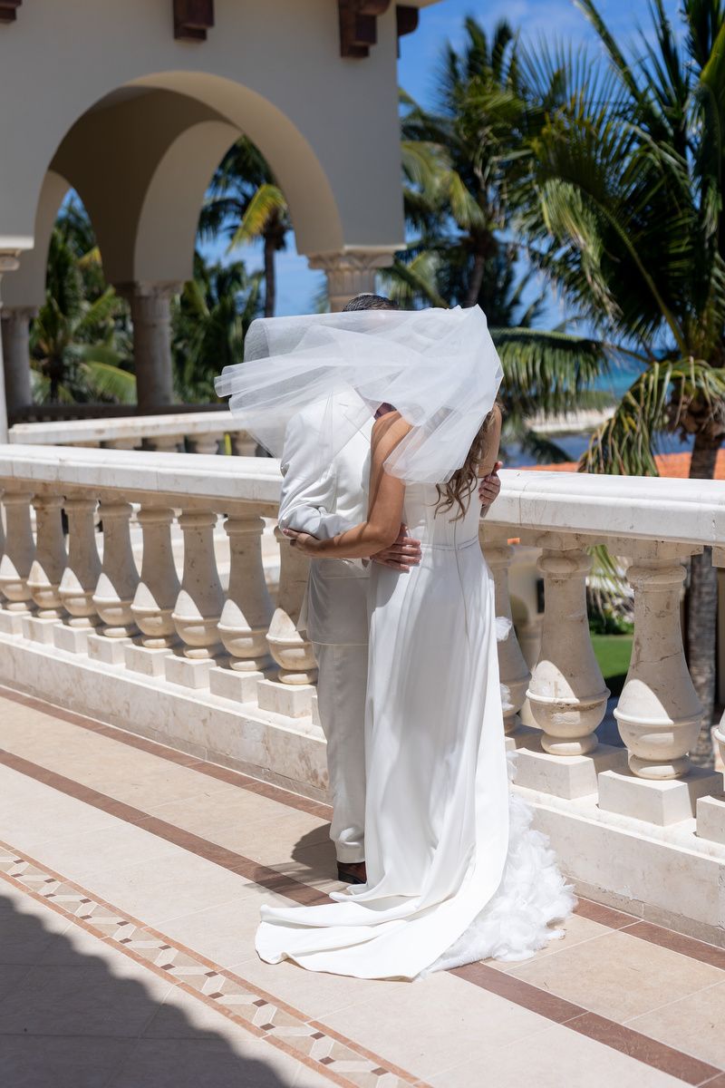 Bride and groom sharing a kiss on a tropical resort balcony, bride in white gown with flowing veil, palm trees in background.