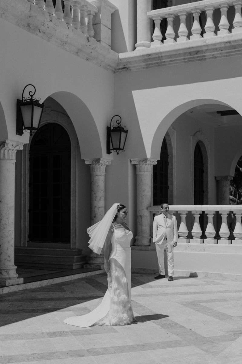 Black and white wedding photo of bride in flowing gown and groom in suit posing at elegant arched colonial venue.