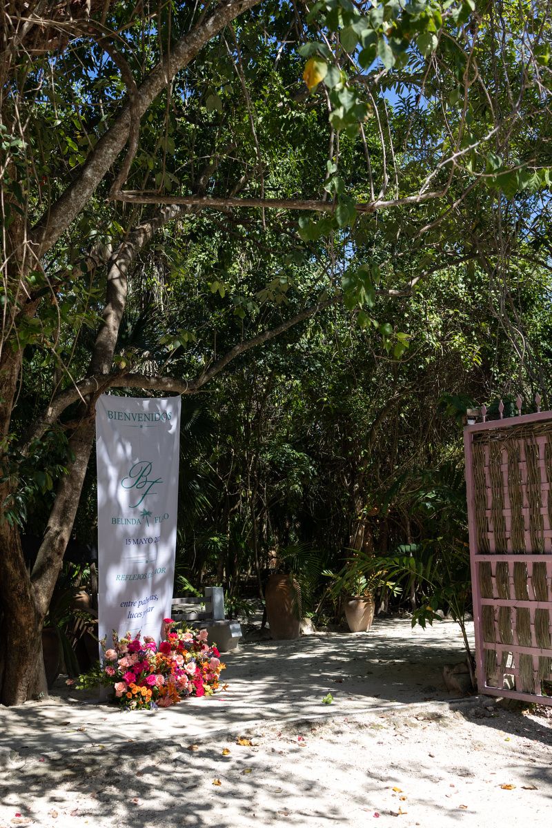 Tropical garden entrance with welcome banner, colorful floral arrangement, and lush green trees lining a sandy pathway.
