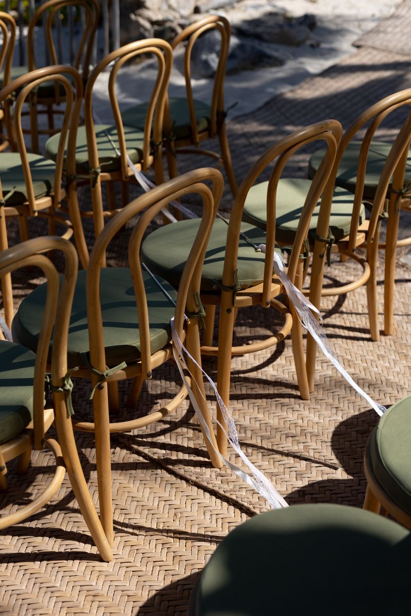 Rows of wooden bentwood chairs with green cushions arranged outdoors on woven flooring, tied with white ribbon.