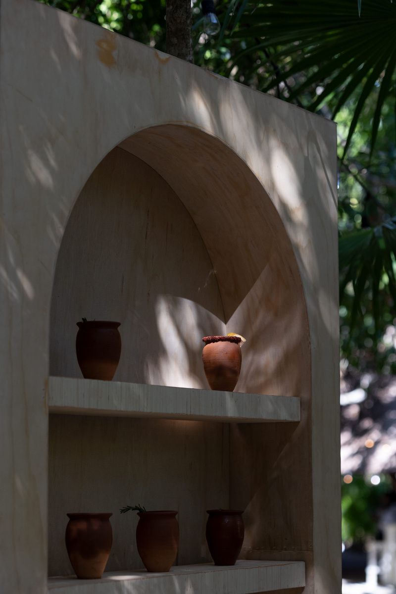 Rustic terracotta clay pots displayed on white stucco shelves with an arched niche, surrounded by lush green tropical foliage.
