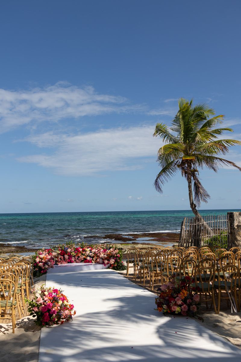 Beachside wedding aisle with white runner, pink floral arrangements, rattan chairs, palm tree, and turquoise ocean under blue sky.