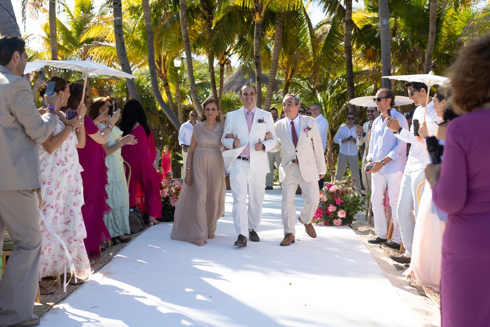 Couple walks down white aisle at tropical outdoor wedding ceremony surrounded by guests in colorful attire and palm trees.