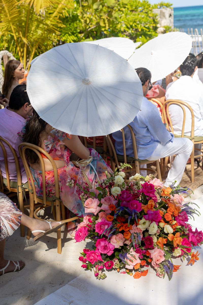 Outdoor wedding ceremony with guests holding white parasols seated near vibrant pink and orange floral arrangements by the beach.