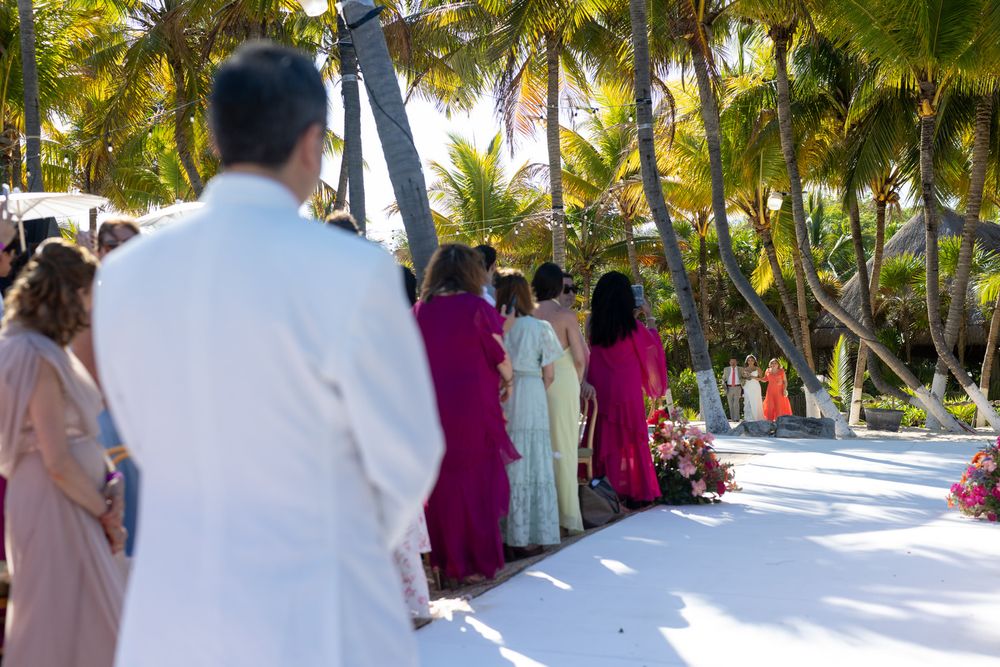 Outdoor beach wedding ceremony with guests in colorful attire standing on white aisle surrounded by tropical palm trees.