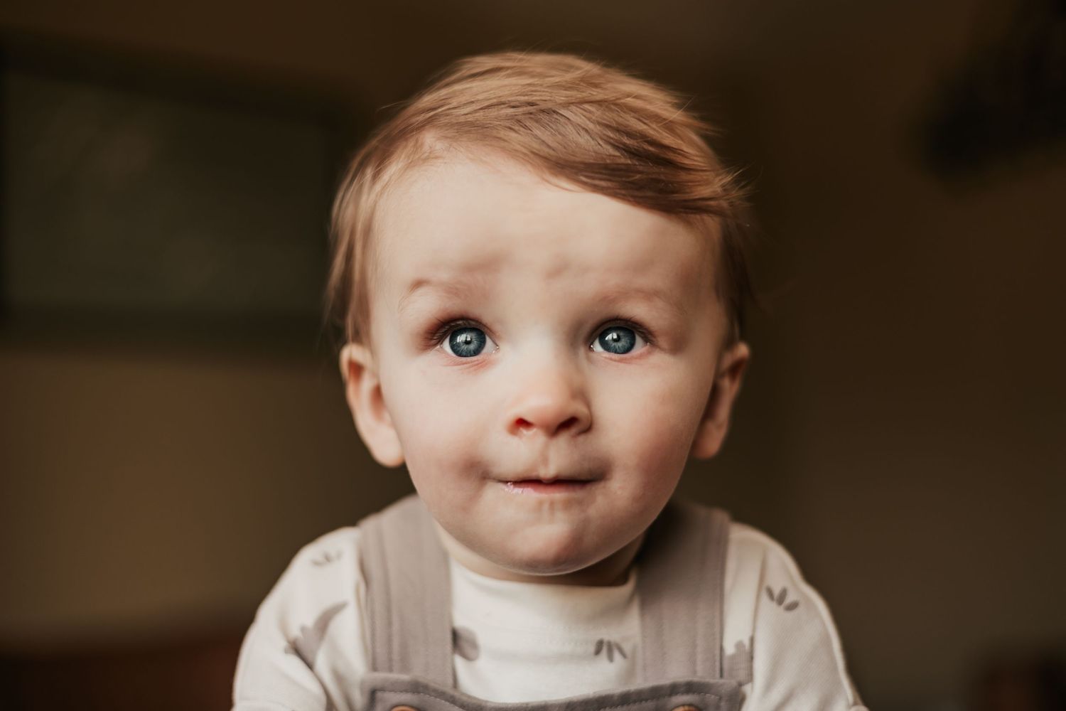 Adorable blue-eyed baby boy with light brown hair wearing gray overalls | Professional Milestone Sessions for Baby