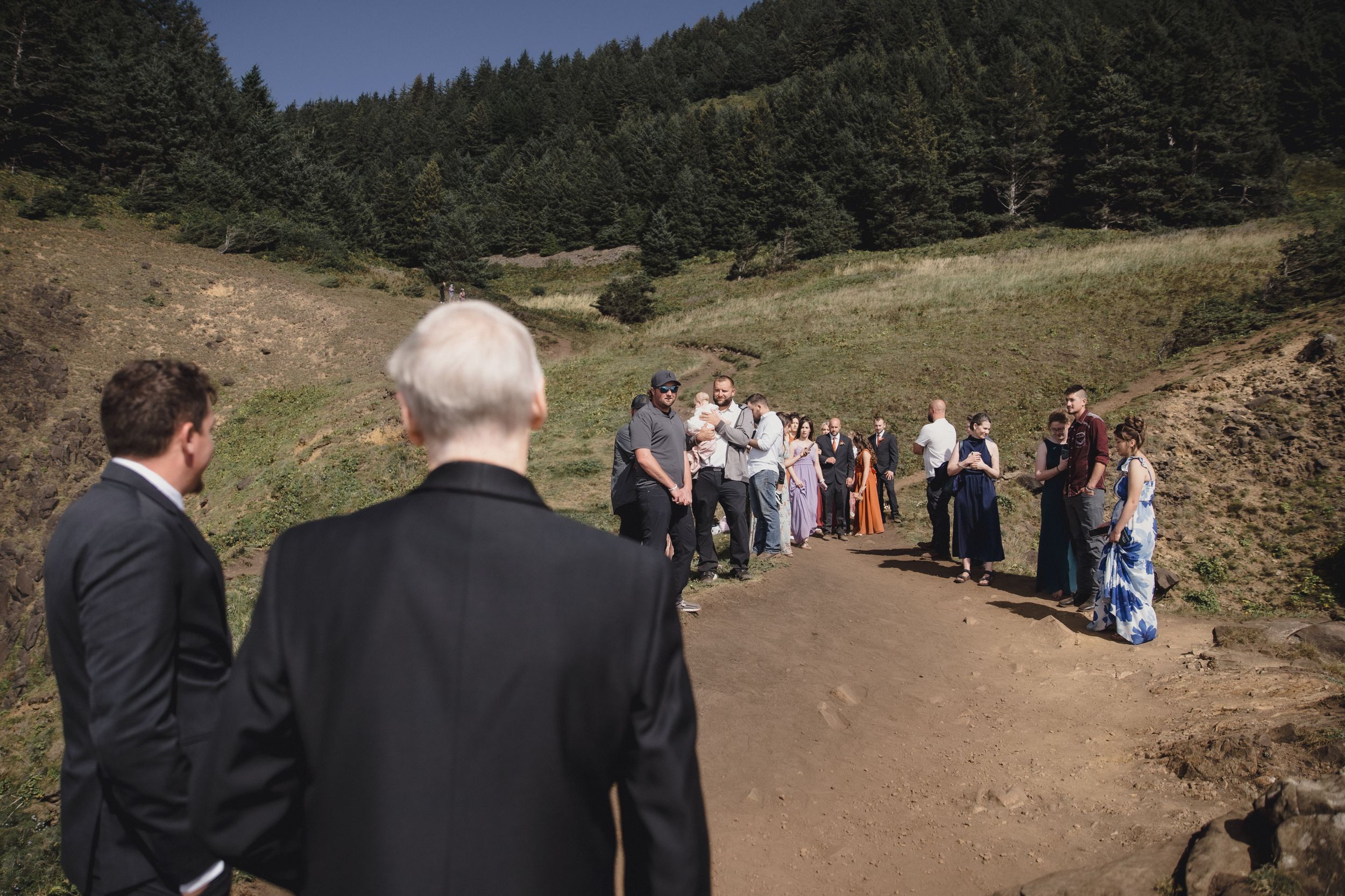 Outdoor wedding ceremony on a rural path surrounded by green hills and pine forest under a clear blue sky.