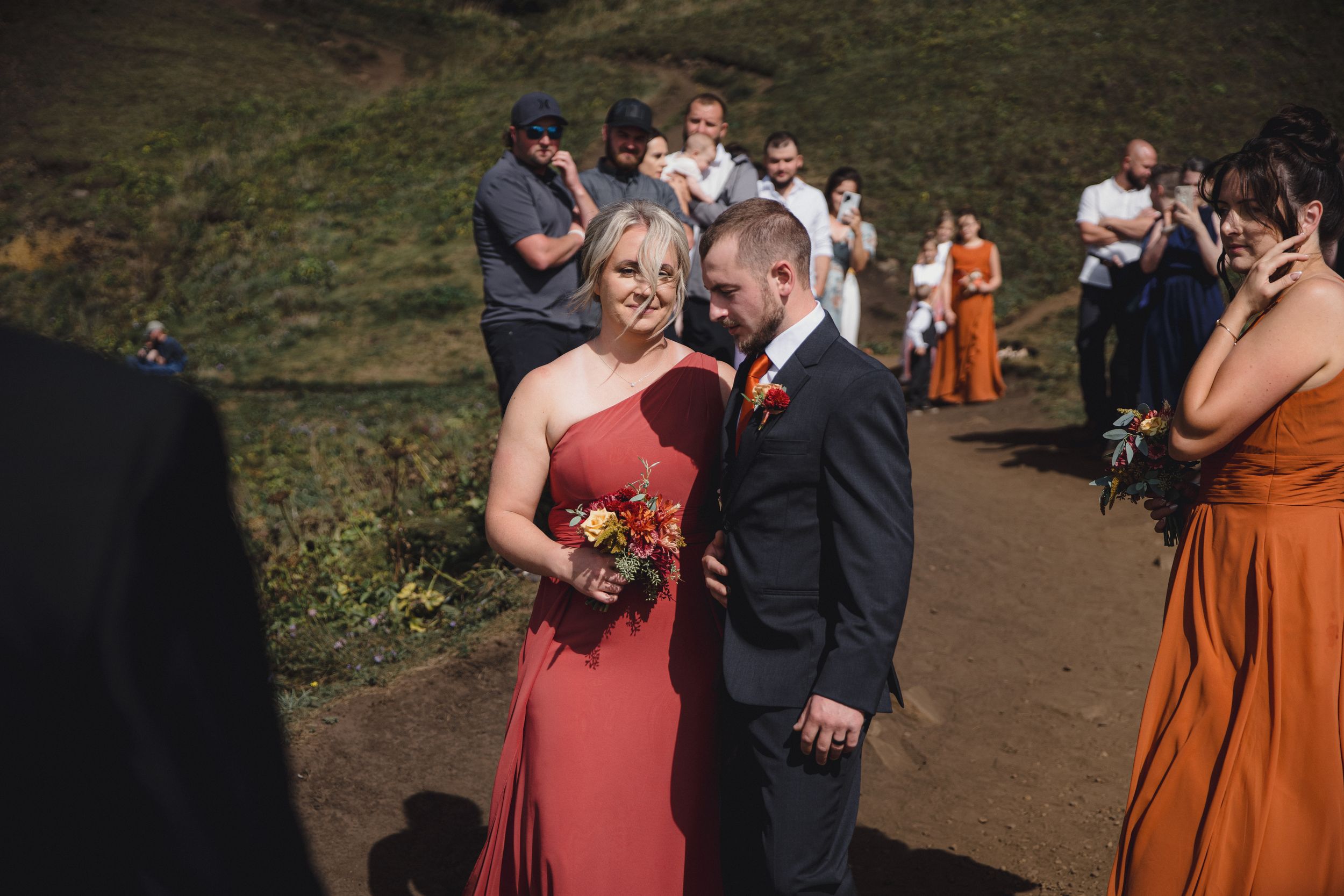 Bride in red dress walks with groom in dark suit at outdoor wedding ceremony, guests watching on hillside.