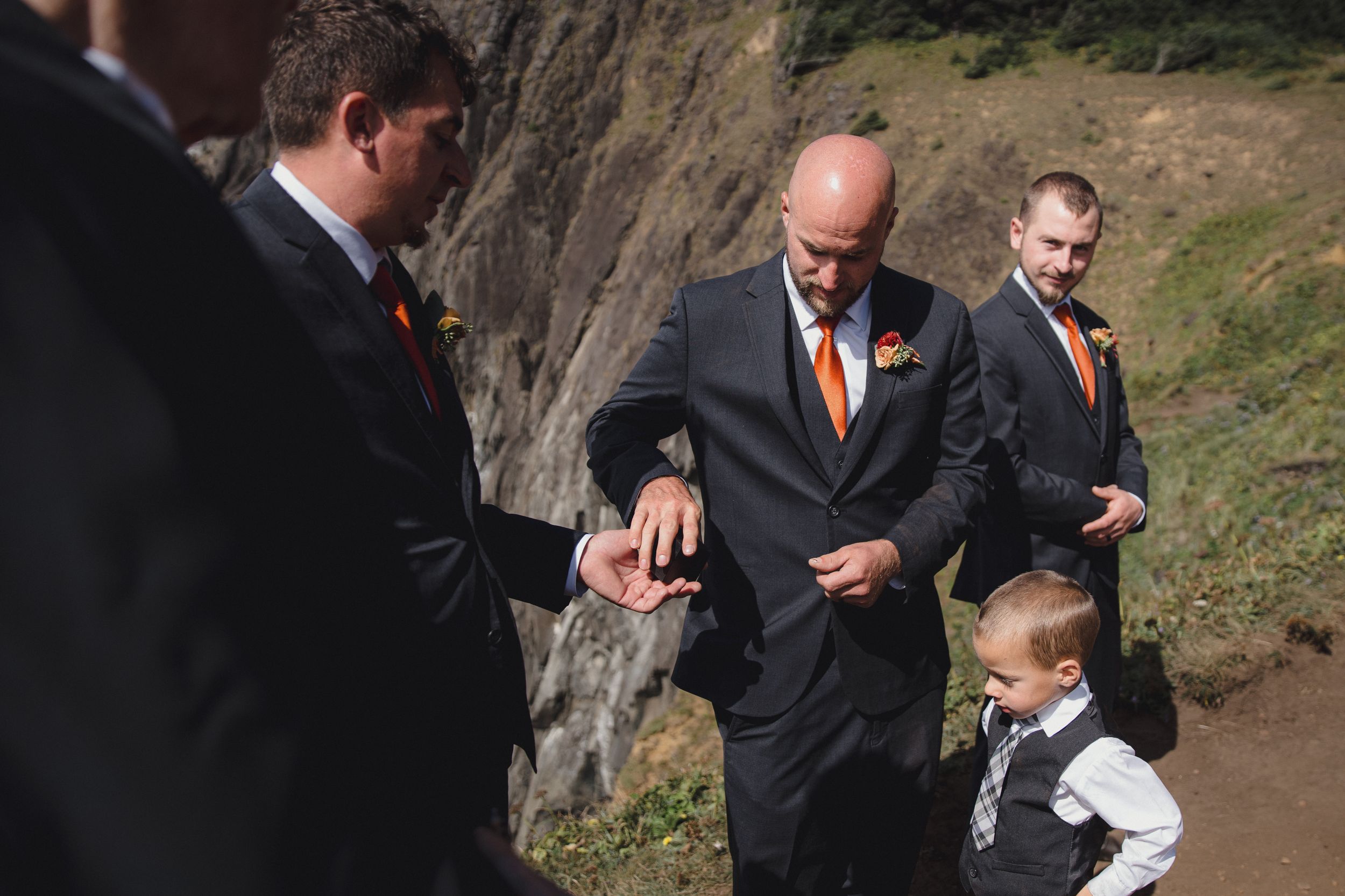 Groom in dark suit with orange tie checks details with groomsmen and ring bearer at outdoor wedding.
