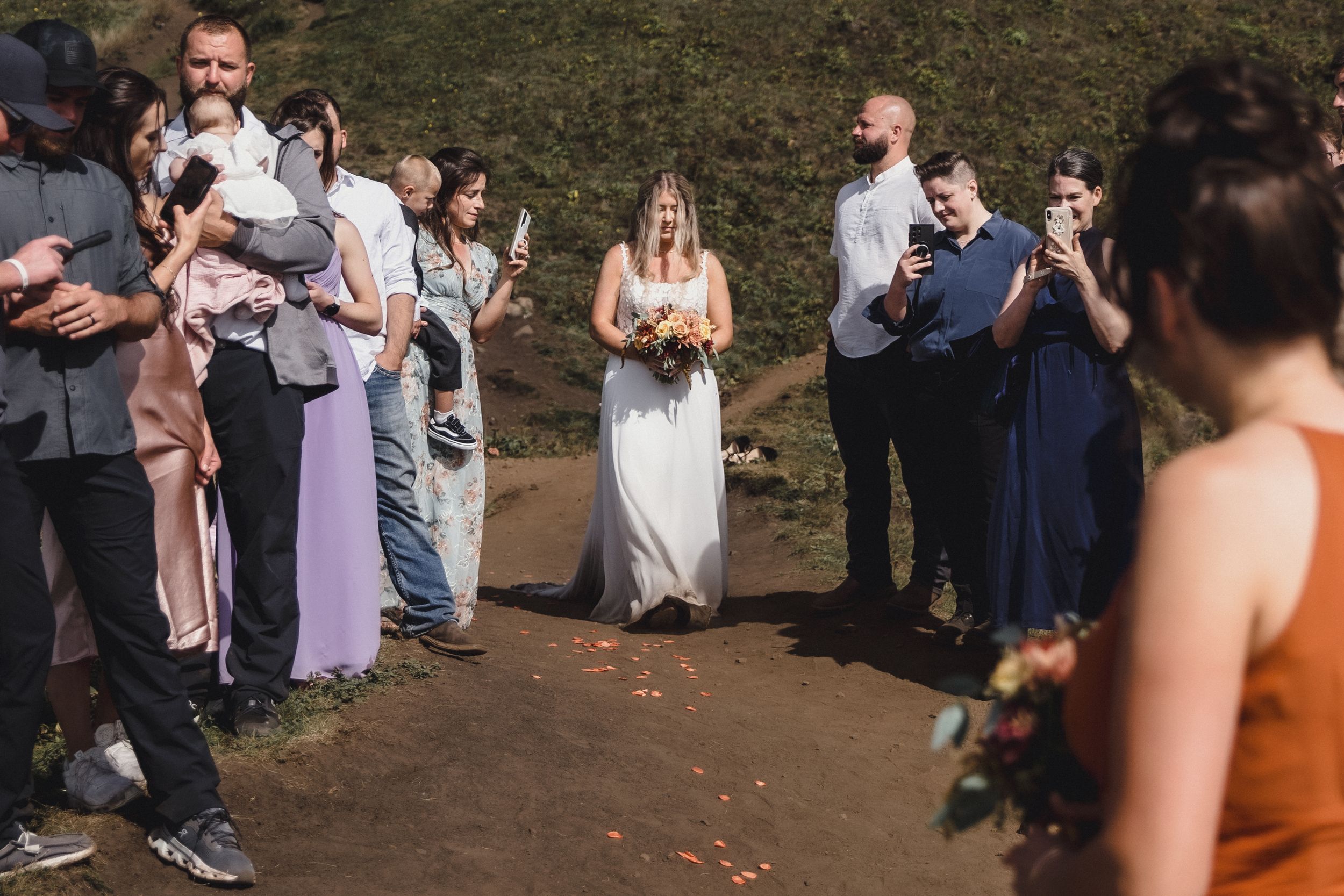 Bride in white gown walks through cheering guests outdoors at a scenic wedding ceremony.