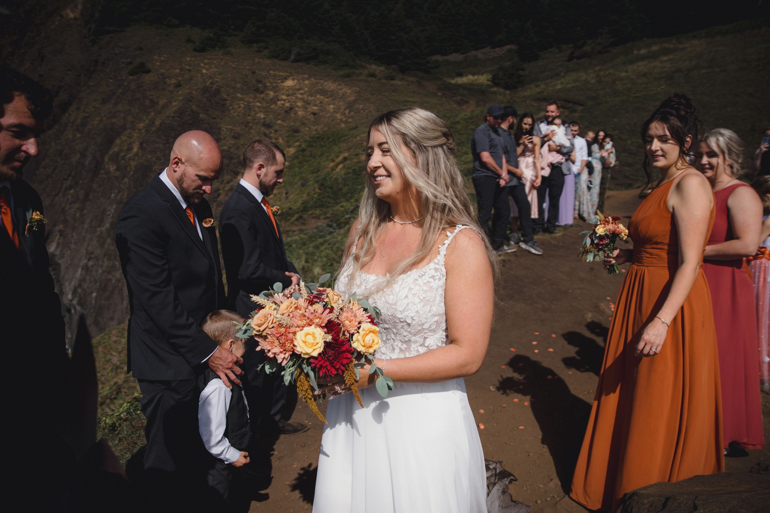 Smiling bride in white lace gown holding colorful bouquet walks with wedding party on rocky outdoor path.